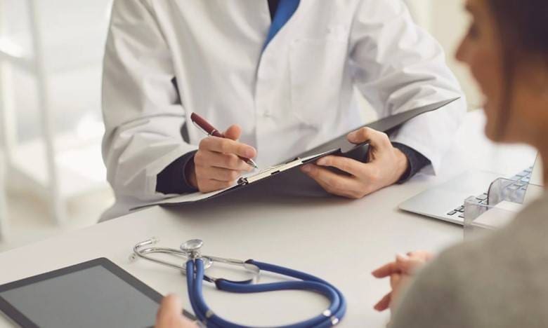 picture of a doctor and patient at a desk- doctor's hands and chart are visible. also visible is the back of a woman's head and profile and a stethoscope and tablet on the table in between them. Doctor is writing in the chart and they appear to be talking to each other.